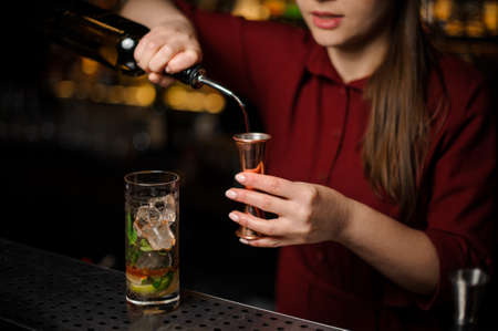 barmaid in a red dress prepares a mojito in a crystal glass, adding dark rum and using bar equipmentの写真素材