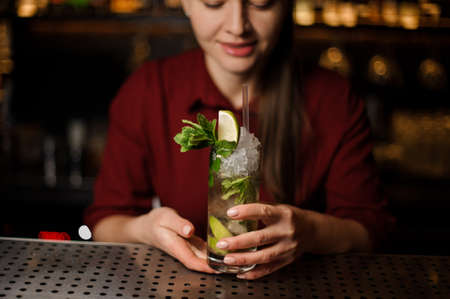 barmaid, a blonde in a red dress, is standing at the bar and holding a ready-made alcoholic cocktail mojito in a glass beakerの写真素材