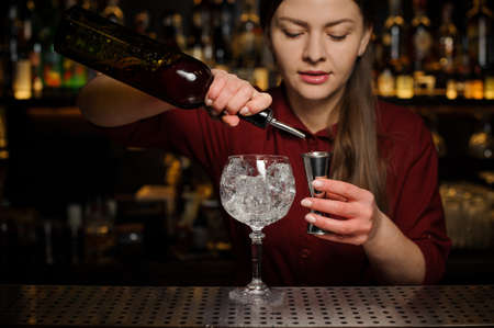 barmaid the blonde in a red dress is standing at the bar and pouring red wine into a glass, preparing an alcoholic cocktailの写真素材