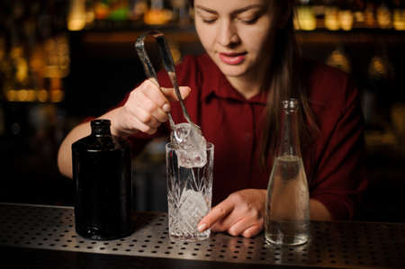 barmaid in a red dress prepares an alcoholic cocktail Caipirinha in a crystal glass, adding ice pieces with ice tongsの写真素材