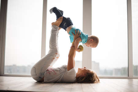 Mother, lying on the floor, holding her little boy, dressed in blue shirt and jeans, up in the air on the background of big windowの写真素材