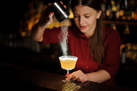 Female bartender serving a fresh yellow cocktail and pouring a spice into it on the bar counterの写真素材