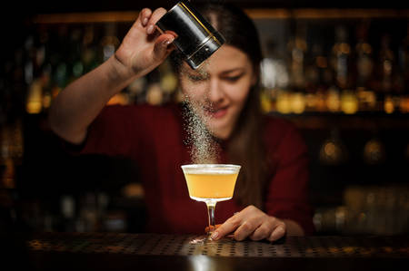 Female bartender serving a fresh yellow cocktail and pouring a spice into it on the bar counterの写真素材