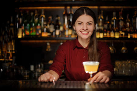 Smiling female bartender serving a fresh delicious yellow cocktail in the beautiful glass on the steel bar counterの写真素材