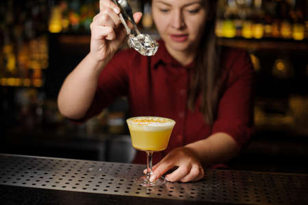 Female bartender adding an ice cube to a fresh delicious yellow cocktail in the beautiful glass on the steel bar counterの写真素材