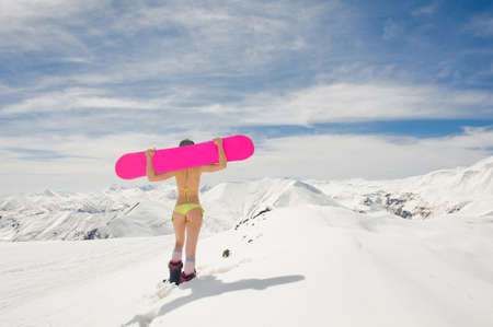 Back view of sexy girl dressed in a bright yellow swimsuit with a snowboard on the shoulders walking in mountainsの写真素材