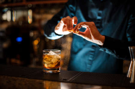 Bartender sprinkling orange peel juice into a cocktail glass filled with a fresh alcoholic drinkの写真素材