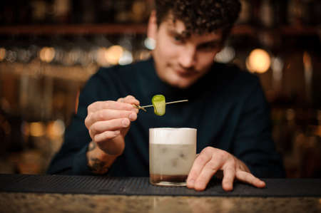 Bartender decorating a fresh cocktail with a fresh cucumber on a wooden skewer on the bar counterの写真素材
