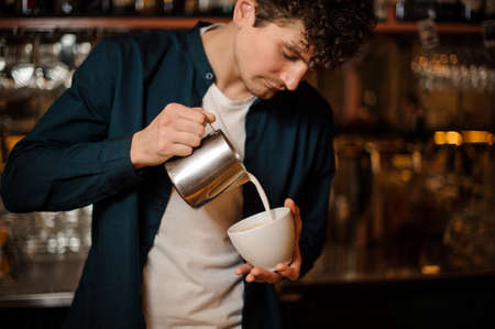 Young curly barman pouring some milk from pitcher into a little white cup in a restaurantの写真素材