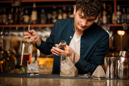 Bartender adding an alcoholic drink from the measuring cup into the glass with ice on the bar counterの写真素材