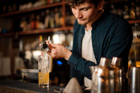 Bartender pouring an alcoholic drink from the measuring cup into the glass with ice on the bar counterの写真素材