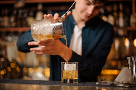 Bartender pouring an alcoholic drink into a glass with ice cube on the bar counterの写真素材