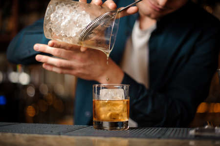Bartender pouring an alcoholic drink from a glass cup into a cocktail glass with a big ice cube on the bar counterの写真素材