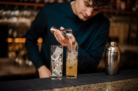 bartender with curly hair and in a blue shirt begins preparing two alcoholic cocktails, pouring alcohol on iceの写真素材