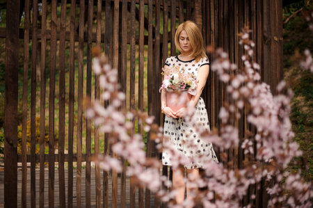 Beautiful blonde girl, dressed in a white black dotted dress, standing near the wooden fence with a...