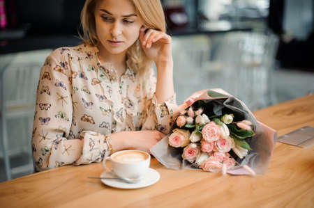 Girl sitting at the table with a cup of coffee and beautiful bouquet of tender rose colour flowersの写真素材