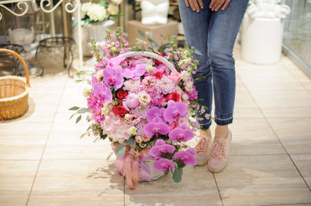 A large wicker basket full of colorful flowers in pink tones near woman legs in a flower shopの写真素材