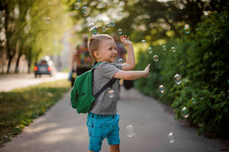 Happy little boy with a backpack playing with soap bubbles in green summer park on sunny dayの写真素材