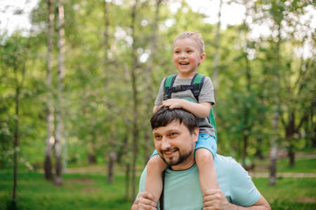 Happy bearded father carrying his son on his shoulders walking in the green summer parkの写真素材
