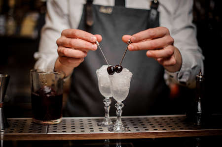 Barman hands decorating two elegant cocktail glasses filled with ice with fresh cherries on the bar counterの写真素材