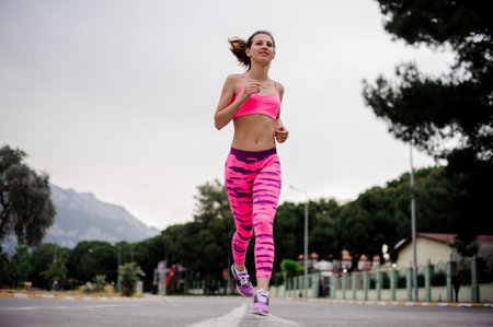 Young and slim woman dressed in pink sportswear running along the highway in the morningの写真素材
