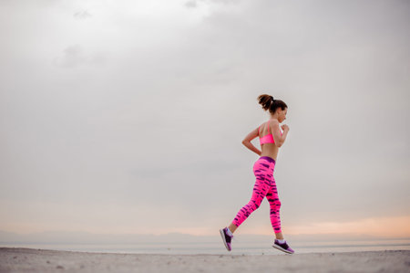 Young beautiful girl dressed in pink sportswear running over the sea coast during the morning sunriseの写真素材