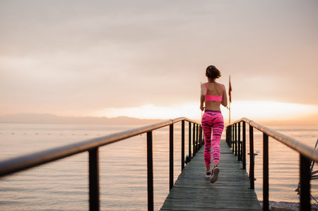 Beautiful woman dressed in pink sportswear running over wooden sea bridge during beautiful morning...