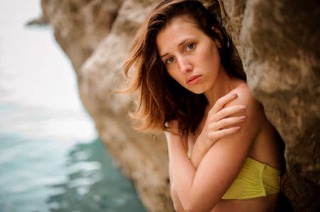 Attractive young redhead woman in yellow bikini swimsuit standing near the rock in the water inside the mountainの写真素材
