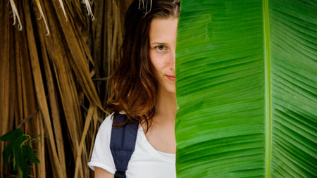 Attractive young redhead girl hiding a half of her face with green leaf on the background of dried leavesの写真素材