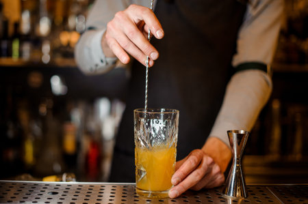 Bartender stirring an alcoholic orange drink in the beautiful glass on the steel bar counter