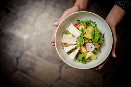 Viev from above girl holding a white bowl with a fresh salad consisting of greenery and brie cheese on the dark blurred backgroundの写真素材