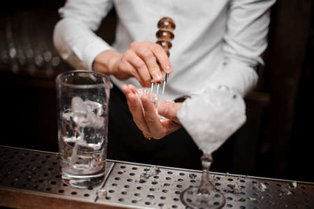 Professional bartender breaking an ice cube with the special tool to prepare a fresh cocktail at the steel bar counterの写真素材