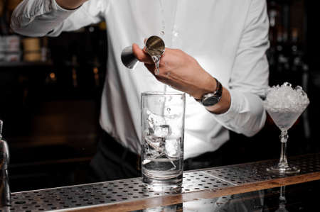 Bartender adding a trasparent alcoholic drink from the measuring cup into the glass with ice on the bar counterの写真素材
