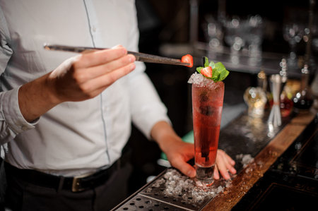 Professional bartender adding a strawberry to a fresh transparent red cocktail with mint leaves at...