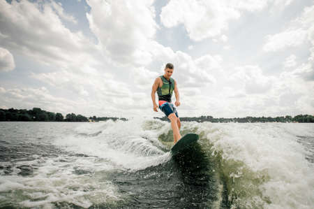 Young attractive brunette man riding on the wakeboard on high wave of motorboat on the lakeの写真素材