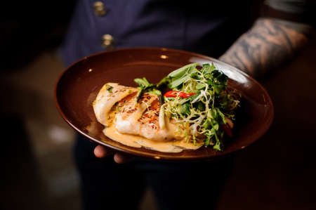 Tattooed bartender holding a plate of roasted chicken fillet and green fresh salad on the one hand on the blurred backgroundの写真素材