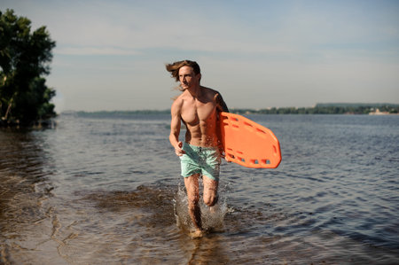 Sexy young long-haired beach lifeguard running along the river bank with life-saving equipment on summer dayの写真素材