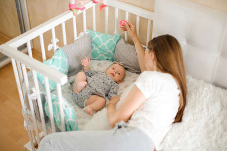 Mother lying on the bed near her little daughter in their cozy domestic room in light colorsの写真素材
