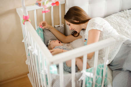 Mother lying on the bed near her little crying daughter in their cozy domestic room in light colorsの写真素材