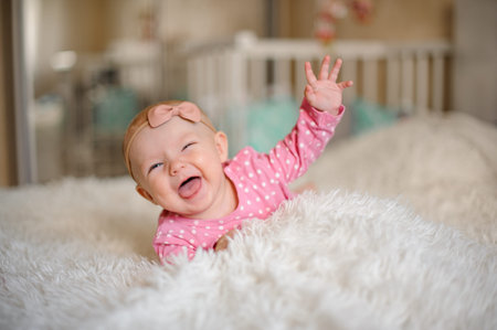Little laughing girl with a bow on her head lying on the white cozy bed in the domestic room