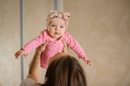 Mother holding her lovely little daughter in the pink romper up in the air in their cozy domestic...