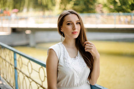 Gorgeous woman with dark hair in elegant white dress standing in the public park on the background of pondの写真素材