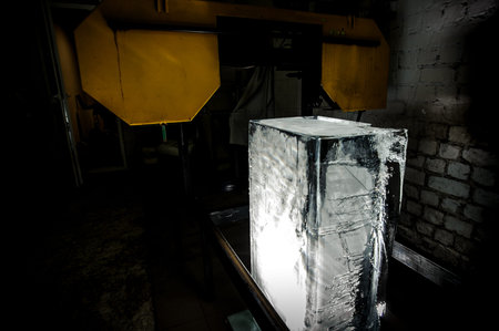 Big clear ice cube standing on the floor under the yellow equipment element on the ice production...