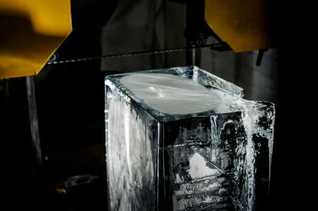 Big clear ice cube standing on the floor under the yellow steel equipment element on the ice production plant in the dark under the light equipment.の写真素材