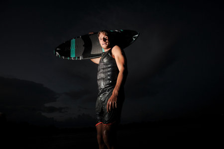 Young man standing with the board on the shoulder in the night on the lake on the background of dark...