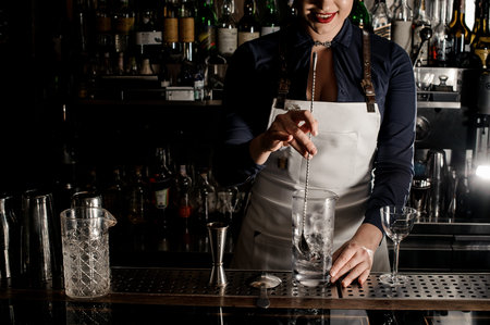 Attractive female bartender in the white apron stirring an alcoholic drink in the glass with ice...