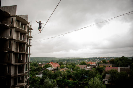 Rear view man making step on the slackline rope on the background of houses among trees and clear...