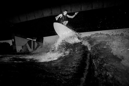 Black and white photo of active and young wakesurfer riding down the river waves against the bridge
