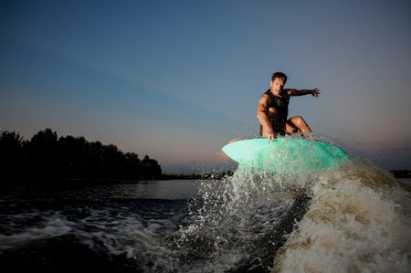 Attractive man riding on the wakesurf down the river on the background of beautiful sky during sunsetの写真素材