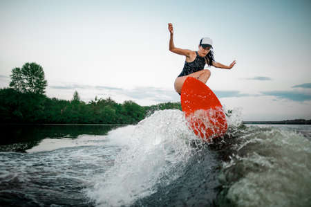 Young attractive brunette woman jumping on the orange wakesurf on high wave of motorboat on the background of treesの写真素材
